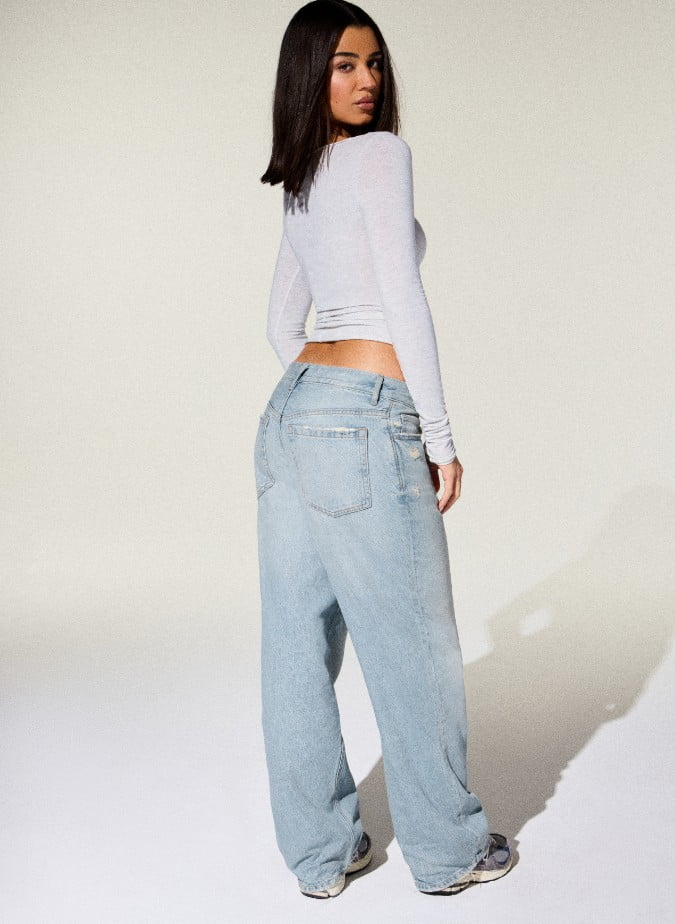 Model poses in a long sleeve light grey shirt and baggy light wash jeans.